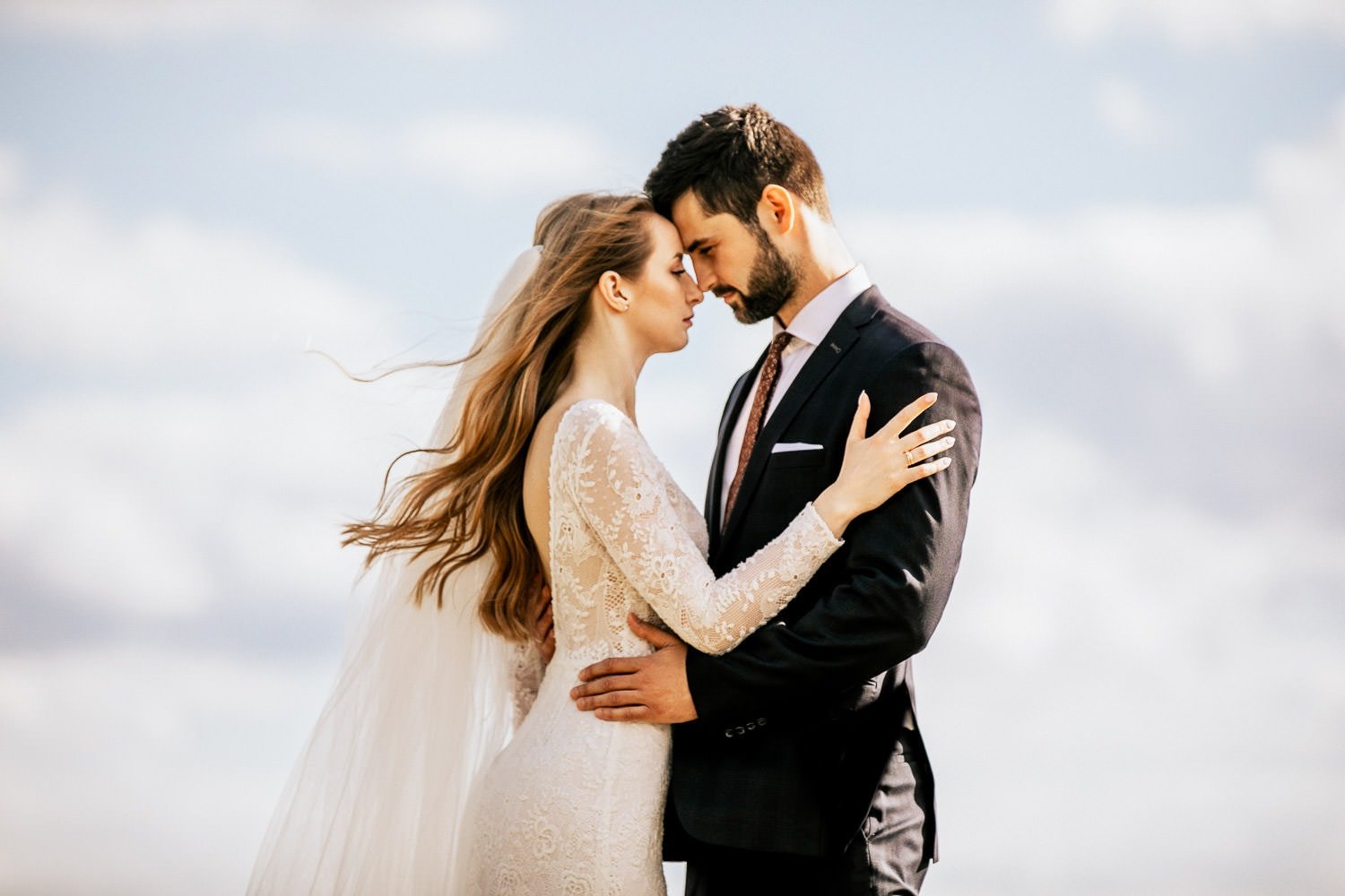 Bride and groom embracing with their foreheads touching beneath an open sky